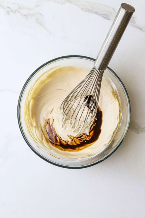 Creamy vanilla frosting with chocolate syrup in a glass mixing bowl with a whisk.
