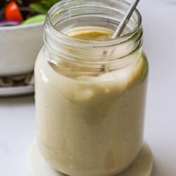 Creamy homemade salad dressing in a glass jar with a spoon, fresh lemon slice, and a side salad in the background.