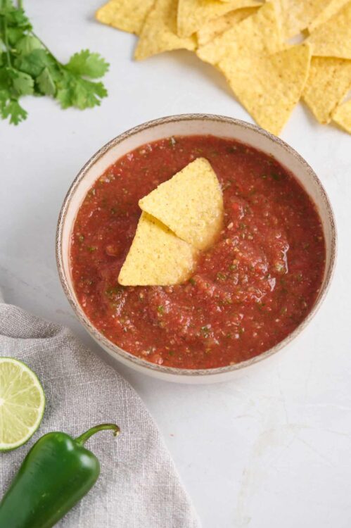 Creamy salsa and tortilla chips in a bowl, fresh lime, and cilantro on white background.