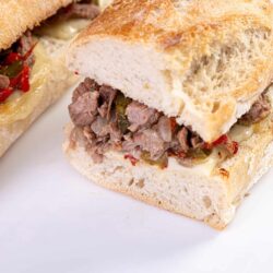 Thinly sliced beef sandwich on a crusty bread roll with melted cheese and roasted peppers on a white plate.