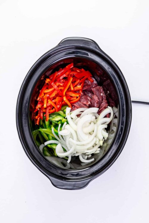 Chopped red bell peppers, green bell peppers, onions, and raw beef in slow cooker for chili recipe.