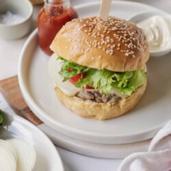 Juicy beef burger with fresh lettuce, tomato, cheese, and toasted sesame bun on white plate.