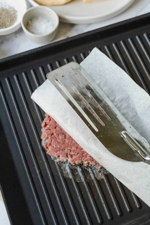Flipped raw burger patty on parchment paper over grill pan, ready for cooking.