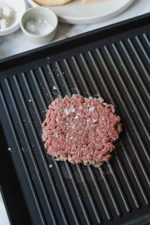 Ground beef patty seasoned on a grill pan, preparing for burgers or grilling.