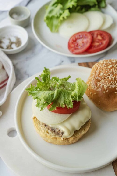 Fresh homemade cheeseburger with lettuce, tomato, onion, melted cheese, and a sesame seed bun on a white plate.