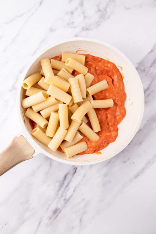 Creamy tomato sauce with uncooked rigatoni pasta in a white skillet on marble surface.