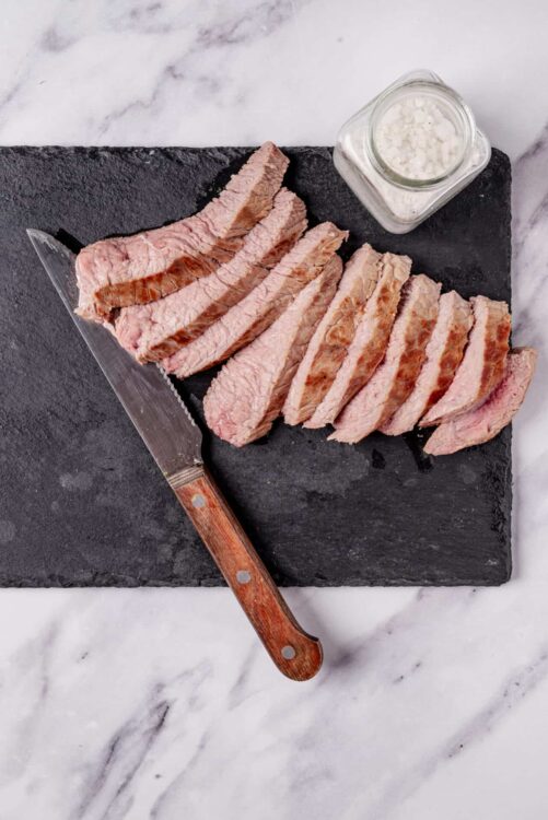 Sliced cooked steak on black cutting board with salt and knife, ready for serving.