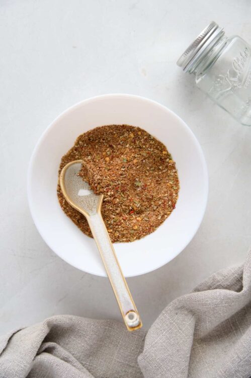 Ground spice blend in a white bowl with a measuring spoon, on a light background.