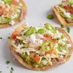 Fresh chicken tostadas topped with guacamole, cheese, tomatoes, and scallions on a white surface.