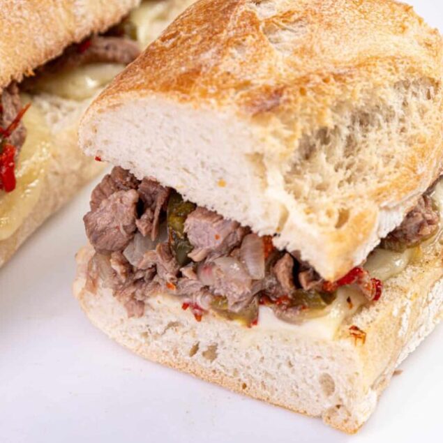 Thinly sliced beef sandwich on a crusty bread roll with melted cheese and roasted peppers on a white plate.