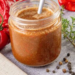 Rich spicy pepper jelly in a glass jar with a metal straw, surrounded by fresh red peppers and black peppercorns.