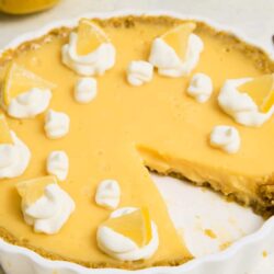 Rich lemon pie with whipped cream and tart lemon wedges in a white ceramic dish.