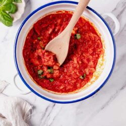 Roasted tomato sauce with fresh herbs in a white enameled pot on marble surface.