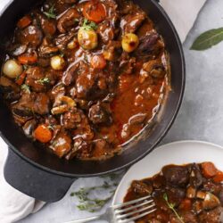 Hearty beef stew with vegetables in a black cast iron skillet, served with a plate of a spoonful of stew on a white dish.