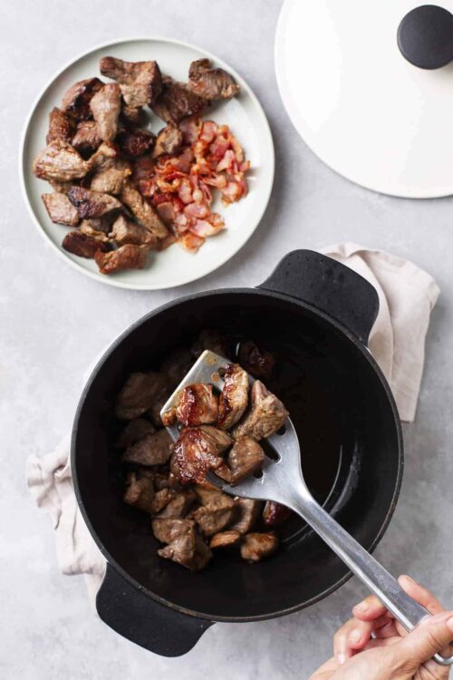 Juicy beef stew meat being transferred from a skillet to a white plate with cooked bacon pieces on top.