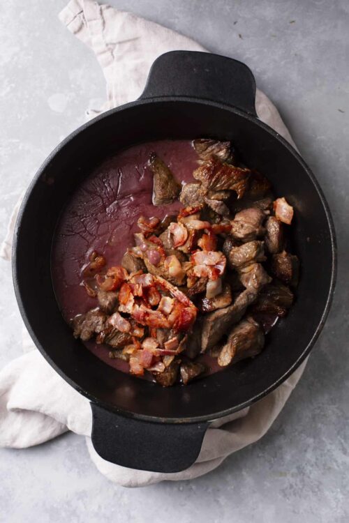 Chunked beef stew meat cooking in a cast iron skillet with bacon bits on top.