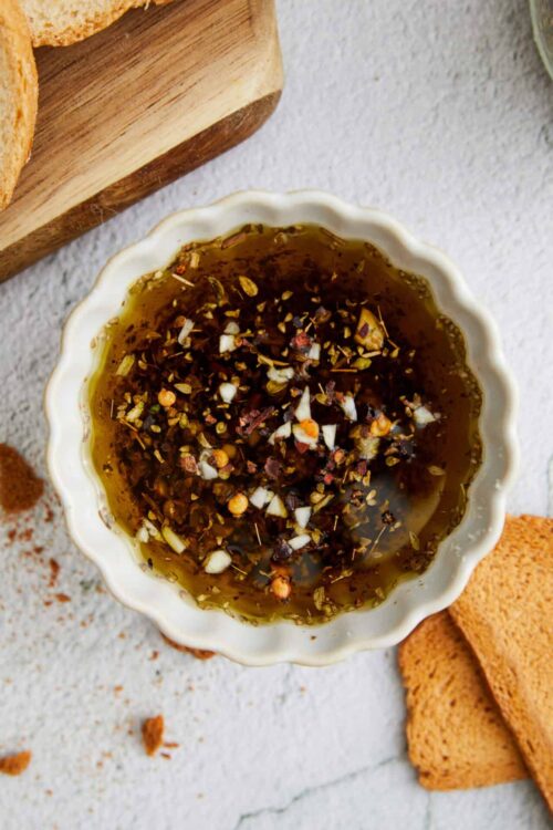 Herb oil and crushed red pepper in a small white bowl on a textured surface with bread slices nearby.