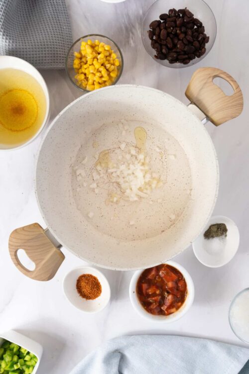 Creamy white onion and garlic mixture in a large mixing bowl with various chopped vegetables and spices around it.