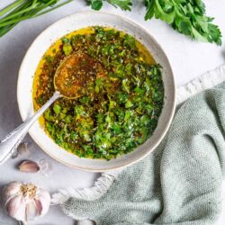 Fresh chimichurri sauce with chopped herbs and olive oil in a bowl with garlic and parsley nearby.
