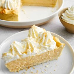 Buttermilk Lemon Pie with whipped topping on a white plate, baked creamy lemon pie dessert with a graham cracker crust, homemade lemon curd filling, and fresh lemon zest garnishes.