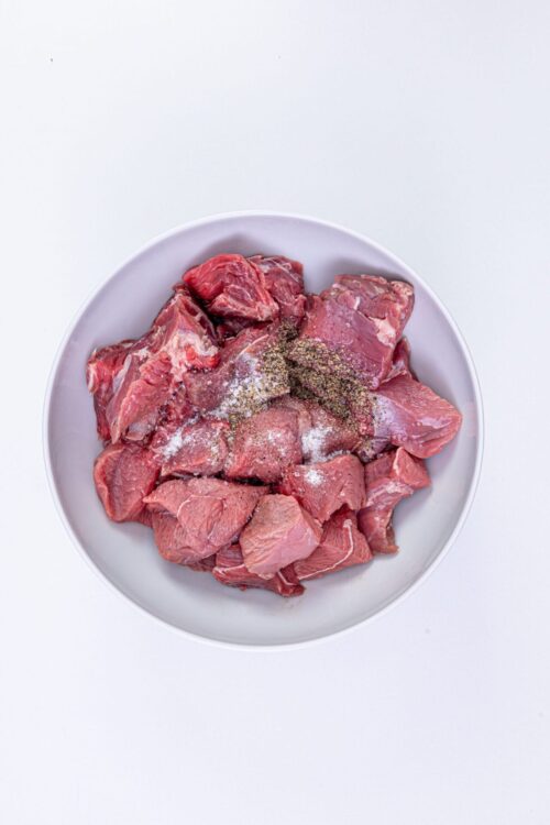 Cubed raw beef with salt, pepper, and spices in a white bowl for cooking or braising.