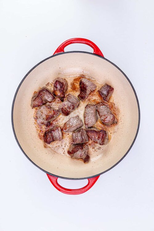 Juicy seared steak cooking in a red Le Creuset cast iron skillet on a white background.