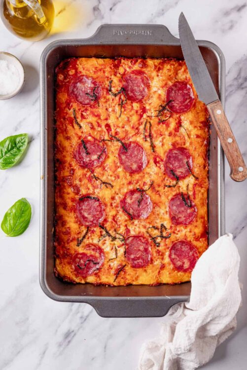 Cheesy pepperoni pizza in a baking dish, fresh basil leaves, and ingredients on a marble countertop.