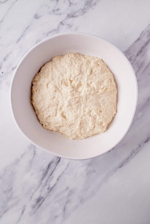 Soft, fluffy pizza dough resting in a white bowl on a marble countertop.