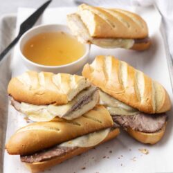 Buttery roast beef baguette sandwich with melted cheese, served with au jus sauce on a white tray.