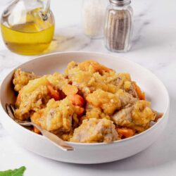 Golden chicken and vegetable pot pie in a white bowl, with salt, pepper, and olive oil on a marble surface.