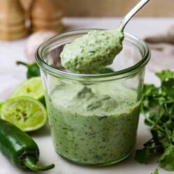 Creamy cilantro jalapeño dressing with lime in glass jar, fresh herbs, and lime slices on marble board.