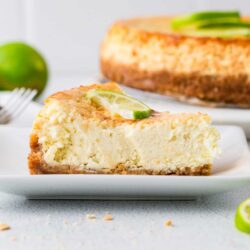 Creamy cheesecake with lime topping on white plate, whole cake in background.