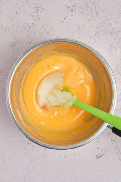 Creamy orange-blended soup in a metal bowl with a green spoon on textured pink background.
