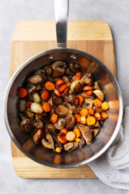Sauteed mushrooms, carrots, and onions in a stainless steel skillet on a wooden cutting board.