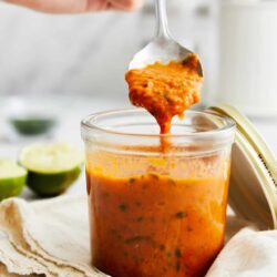 Savory chicken enchilada soup in glass jar with spoon, fresh lime slices in background.