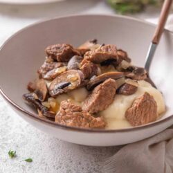 Succulent beef stew with mushrooms over mashed potatoes in a white bowl, cozy home-cooked comfort food, browned meat, savory recipe.