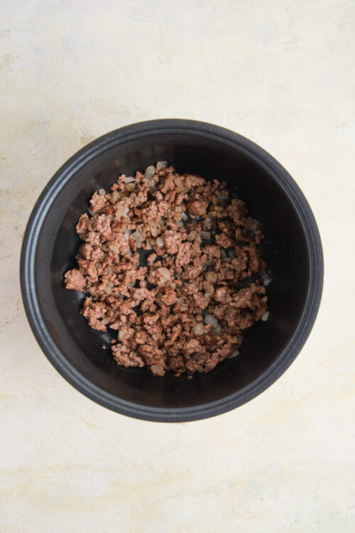 Ground beef and diced onions cooking in a black slow cooker.