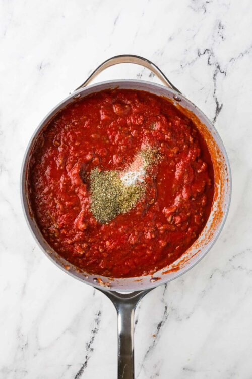 Rich homemade marinara sauce in a stainless steel pot with herbs, on a white marble surface.
