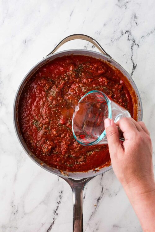 Rich homemade marinara sauce simmering in a skillet with a hand pouring in water for added flavor, perfect for pasta or Italian dishes.