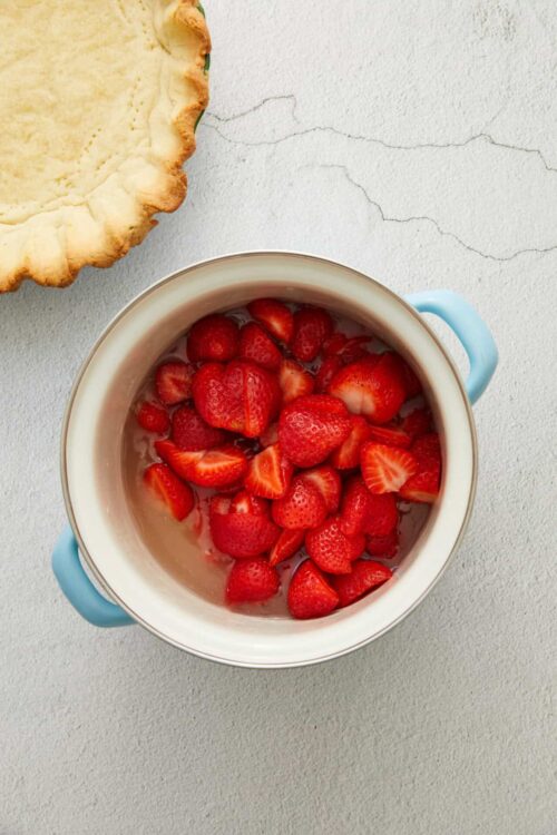 Fresh strawberry slices in a white bowl with blue handles on a white textured surface, perfect for baking and dessert recipes.