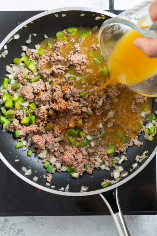 Ground beef and chopped green onions cooking in a skillet with broth being poured in.