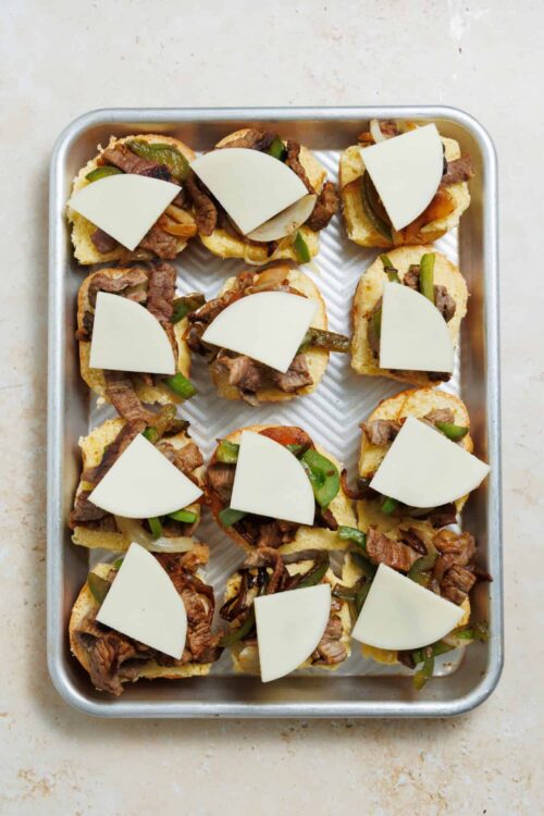Sliced beef and pepper open-faced sandwiches with cheese on baking sheet.