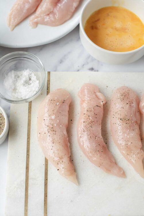 Fresh raw chicken breasts seasoned with salt and pepper on a cutting board.