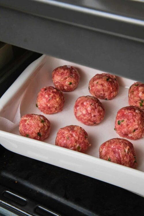 Juicy meatballs baking in oven on white tray.