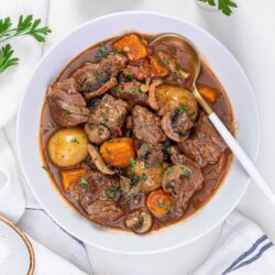 Savory beef stew with carrots and potatoes in a rich broth, served in a white bowl, garnished with chopped herbs.