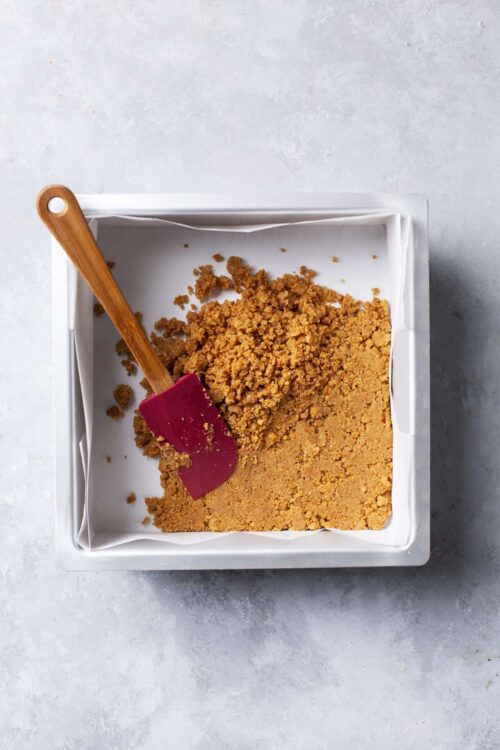 Crushed graham crackers in a white baking dish with a red spatula for s'mores crust.