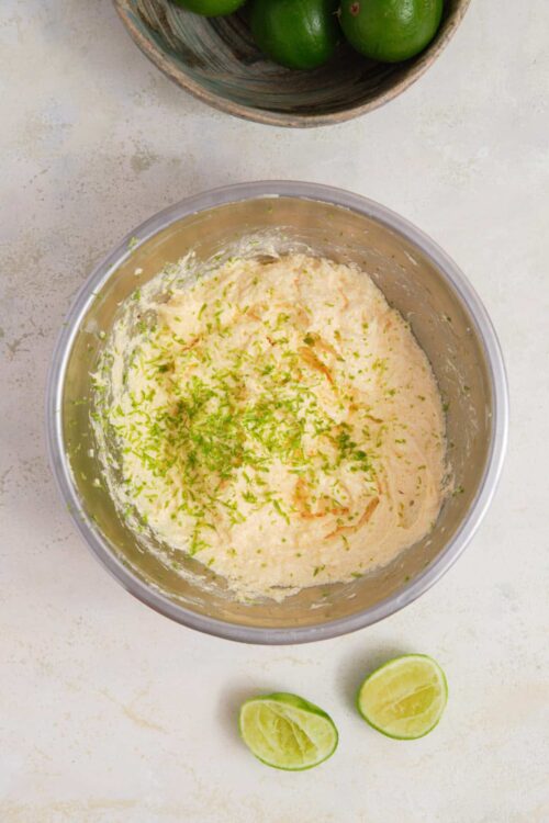 Lime-zested creamy mixture in a metal bowl with lime wedges and a bowl of limes nearby.