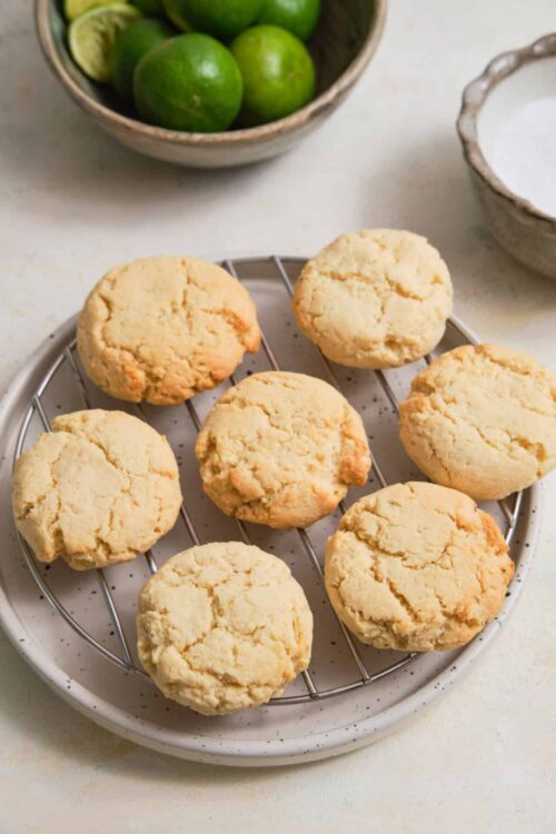 Buttery lemon cookies on cooling rack with limes in background.