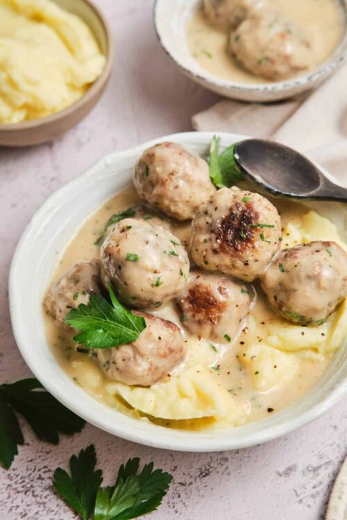 Creamy Swedish meatballs served over mashed potatoes with parsley garnish in a white bowl.