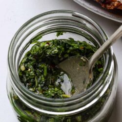 Fresh herb chimichurri sauce in a glass jar with a spoon, ideal for grilled meats and vegetables.
